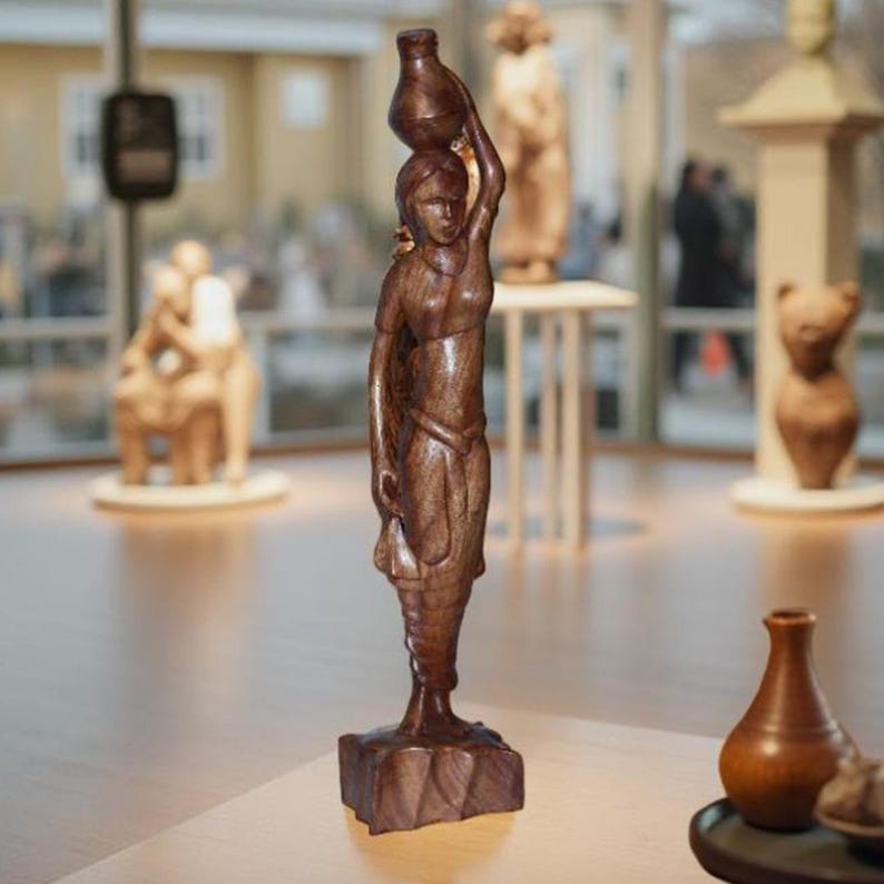 May include: A detailed wooden sculpture of a woman carrying a water jug on her head. The figure is carved from dark brown wood and stands on a square base. Other wooden sculptures are visible in the background, suggesting a collection of art.