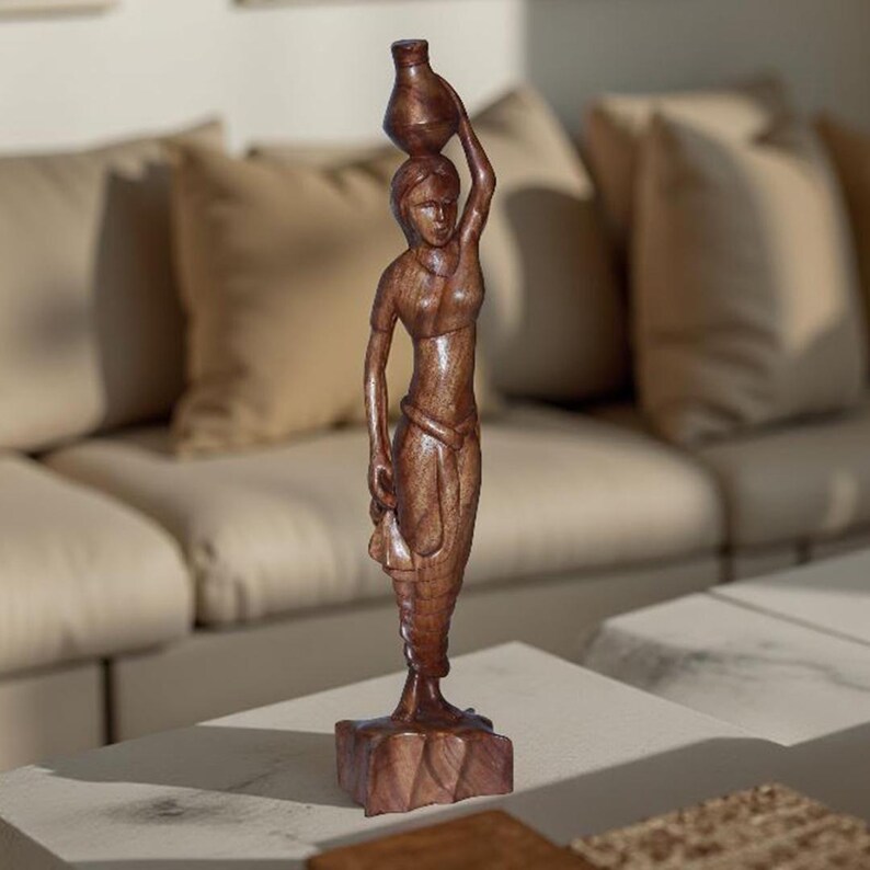 May include: A brown wooden sculpture of a woman carrying a vase on her head. The figure is carved in detail, with flowing garments and a textured base. The sculpture is displayed on a white surface.