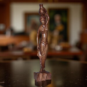 May include: A carved wooden sculpture of a woman carrying a jug on her head. The figure is detailed with flowing garments and stands on a square base. The wood has a rich, dark brown color, and the sculpture is set against a blurred background.