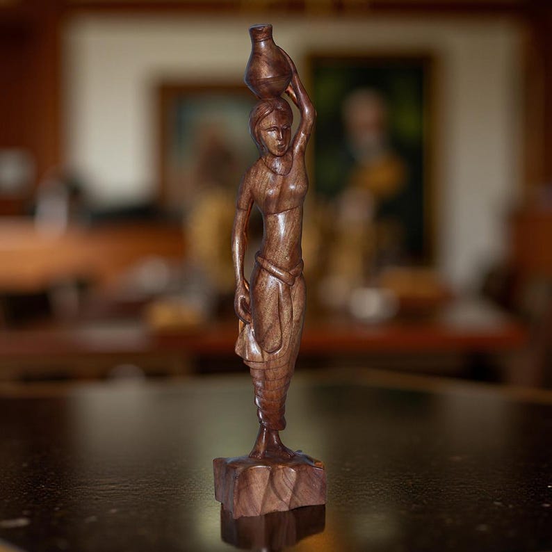 May include: A carved wooden sculpture of a woman carrying a jug on her head. The figure is detailed with flowing garments and stands on a square base. The wood has a rich, dark brown color, and the sculpture is set against a blurred background.