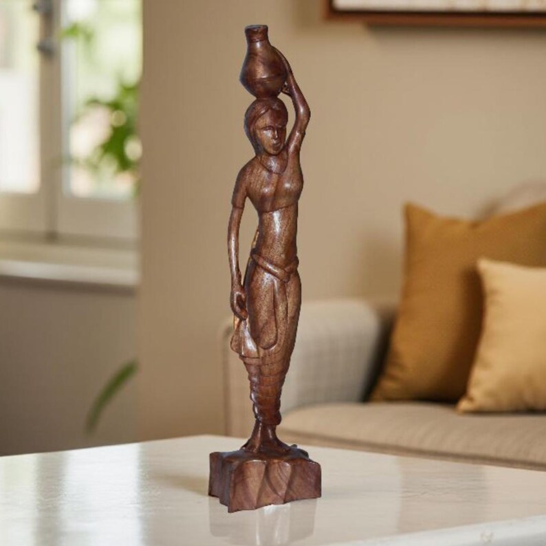 May include: A brown wooden sculpture of a woman carrying a vase on her head. The figure is carved with detailed features and stands on a square base. The sculpture is a decorative item.