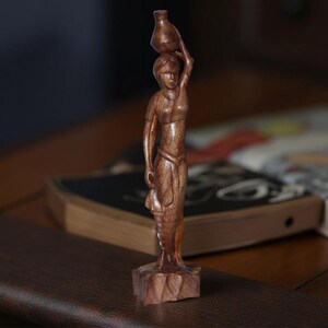 May include: A wooden figurine of a woman carrying a jug on her head. The figure is carved from a light brown wood and stands on a square base. The woman is depicted in a flowing garment, with her arm raised to support the jug. The figurine is a decorative item.