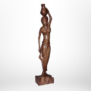 May include: A wooden sculpture of a woman carrying a water jug on her head. The figure is carved from a dark brown wood and stands on a square base. The woman is depicted in a flowing garment.