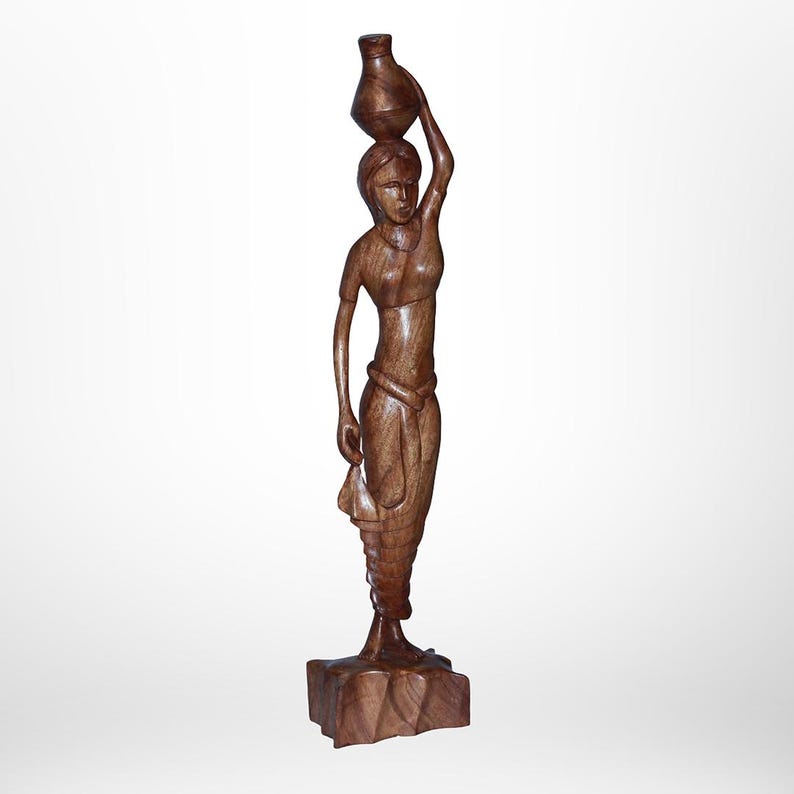 May include: A wooden sculpture of a woman carrying a water jug on her head. The figure is carved from a dark brown wood and stands on a square base. The woman is depicted in a flowing garment.