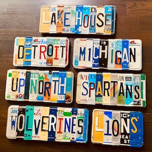 May include: A set of 8 vintage license plates with different city and state names, including Detroit, Michigan, Up North, Spartans, Wolverines, and L10NS. The plates are arranged in two rows of four, with the top row featuring the names of cities and states, and the bottom row featuring the names of teams or organizations.