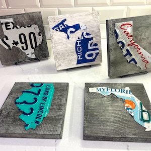 May include: Five wooden plaques with state outlines made from repurposed license plates. The states are Texas, Michigan, California, Kentucky, and Florida. The plaques are painted in shades of gray and blue.