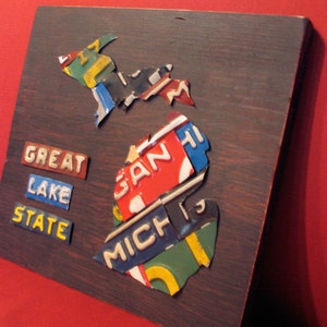 May include: A wooden plaque shaped like the state of Michigan, decorated with vintage license plates. The plates spell out "MICH", "GAN", and "GREAT LAKE STATE".