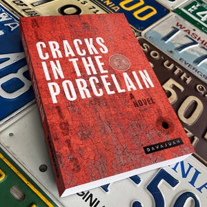 May include: A hardcover book titled "Cracks in the Porcelain: A Novel" with a red cover featuring white text and a cracked texture. The book has a circular emblem and the author's name, Davajuan, is printed at the bottom.