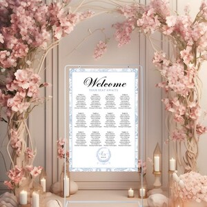 May include: A wedding seating chart with a floral design. The sign reads "Welcome, Your Seat Awaits" and lists guest names by table. The sign is framed by pink floral arrangements and candles.