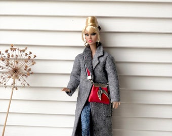 May include: A doll wearing a gray coat, blue jeans, and red handbag with a gold chain. The doll has blonde hair styled in a bouffant and is wearing gold hoop earrings. A gold dachshund figurine is in the foreground.