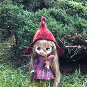 May include: A doll with long blonde hair wearing a red and orange knitted hat and a purple dress with floral print. The doll is holding a small branch with green leaves. The doll is standing in a grassy area.
