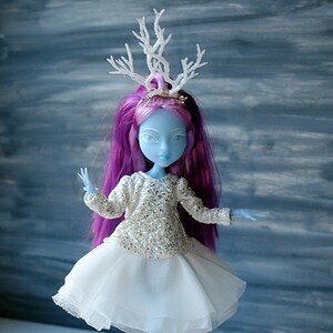 May include: A blue doll with long purple hair wearing a white sparkly top and a white tulle skirt. The doll is wearing a white antler headband.