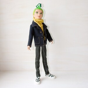 May include: A doll with green hair and a green lizard on its head. The doll is wearing a dark blue jacket with a yellow scarf, black trousers, and black and white chequered shoes.
