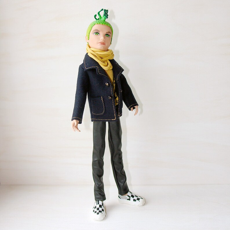 May include: A doll with green hair and a green lizard on its head. The doll is wearing a dark blue jacket with a yellow scarf, black trousers, and black and white chequered shoes.