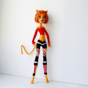 May include: A doll with orange and black striped hair, wearing a red top and black and white checkered leggings with a colorful pattern. The doll has a tail and is posed with one leg slightly bent.