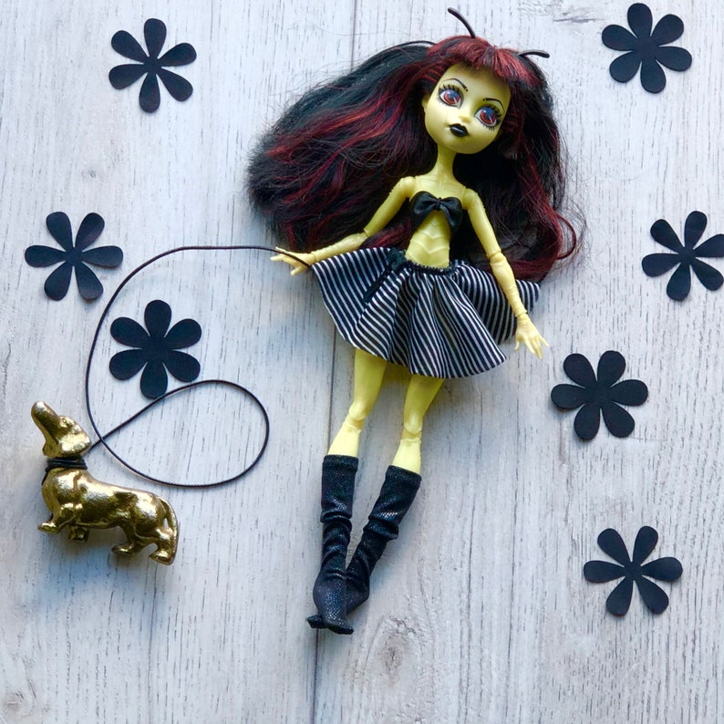 May include: A yellow doll with black and white striped skirt and black boots, holding a leash attached to a gold dachshund figurine. The doll has black and red hair and is wearing a black bow. The background is a light wood surface with black paper flower cutouts.