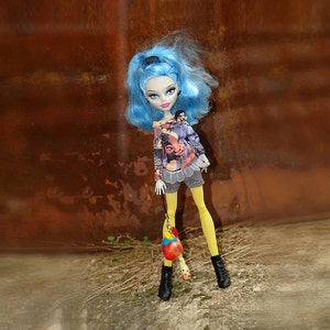 May include: A fashion doll with blue hair, wearing a patterned top, gray shorts, yellow tights, and black boots. The doll is holding a colorful, round accessory. The background is a rusty brown metal sheet.