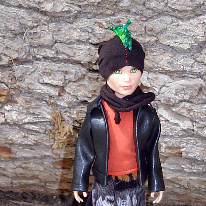 Puede incluir: Una mu&ntilde;eca que lleva una chaqueta de cuero negra, un gorro negro y verde con un dise&ntilde;o verde puntiagudo y una bufanda negra. La mu&ntilde;eca tiene ojos verdes y lleva una camisa roja y naranja y pantalones negros y grises.