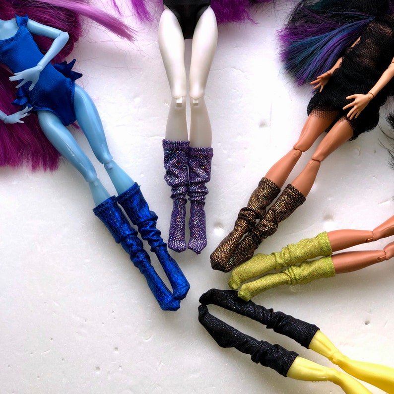 May include: Five pairs of doll boots in various colors and styles. The boots are made of fabric and have glitter accents. The boots are arranged in a circle on a white surface.