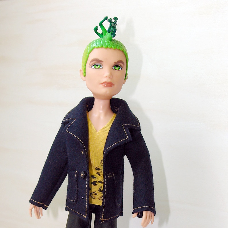 May include: A doll with green hair and a blue jacket. The doll has green eyes and is wearing a yellow shirt.