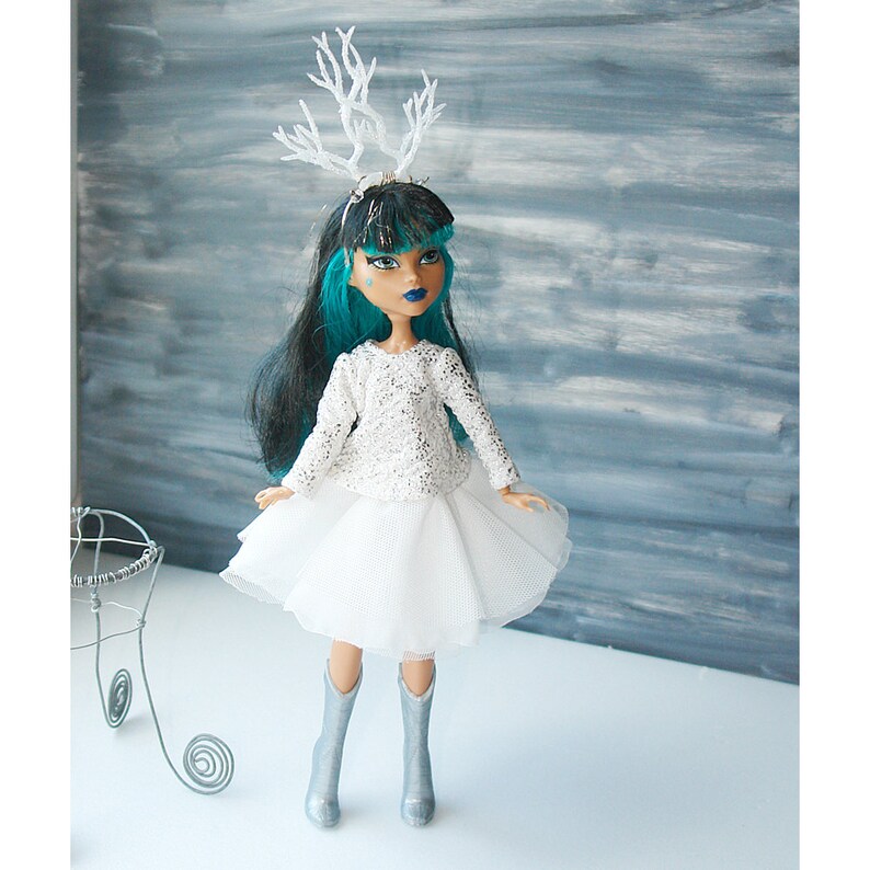 May include: A doll with long black hair and teal blue hair, wearing a white sparkly top, a white tulle skirt, silver boots, and a white antler headband.
