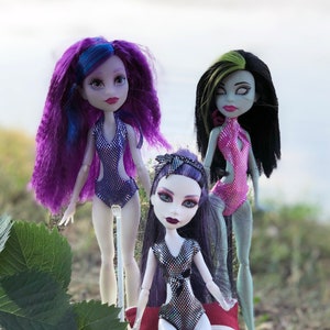 Pode incluir: Três bonecas de moda usando roupas de banho coloridas. A boneca à esquerda tem cabelo roxo longo e está usando um maiô azul brilhante. A boneca do meio tem cabelo roxo longo e está usando um maiô prateado brilhante. A boneca à direita tem cabelo preto e verde longo e está usando um maiô rosa.