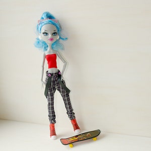 May include: A blue-haired doll with red and black clothing and a skateboard. The doll has a red top, black and white plaid pants, and black suspenders. The skateboard is black and yellow with a graphic design.