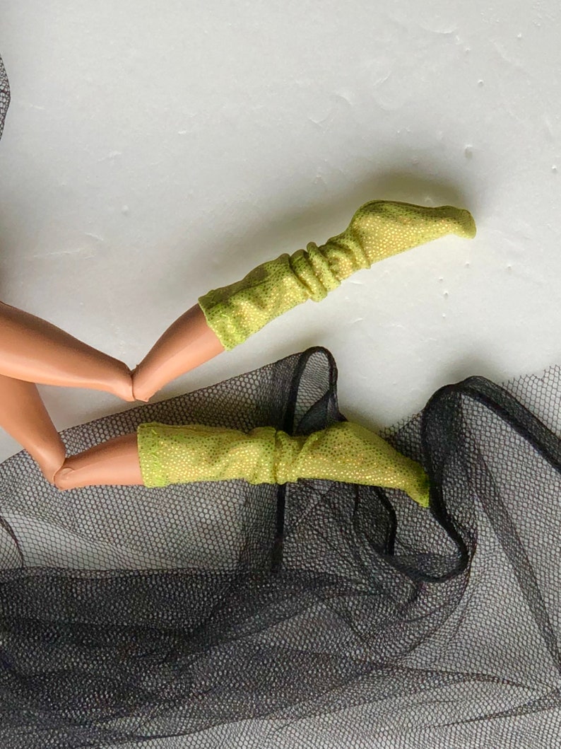 May include: A pair of lime green sparkly doll socks with a ribbed texture. The socks are on a black mesh fabric.