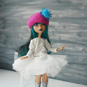 May include: A doll with teal hair wearing a pink beret with a blue pom-pom, a white sparkly top, and a white tulle skirt.