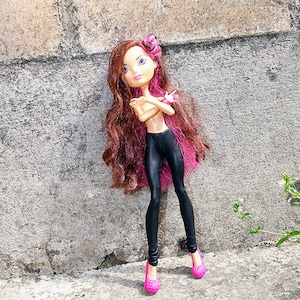 May include: A doll with long pink and brown hair, wearing black leggings and pink shoes. She is standing with her arms crossed in front of a gray stone wall.
