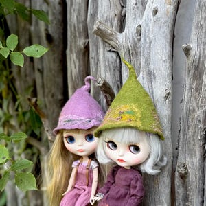 May include: Two dolls wearing whimsical felt hats. One doll has a lavender hat with a lace trim, and the other has a green hat with decorative embellishments. Both dolls have large eyes and are dressed in purple and pink outfits.