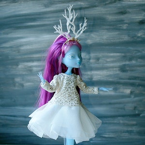 May include: A blue doll with pink hair wearing a white tulle skirt and a sparkly silver top. The doll is wearing a white antler headband.