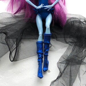 May include: A blue and black doll with long purple hair wearing a blue one-piece swimsuit and blue boots. The doll is posed with one leg bent and the other leg straight. The doll is sitting on a black tulle fabric.