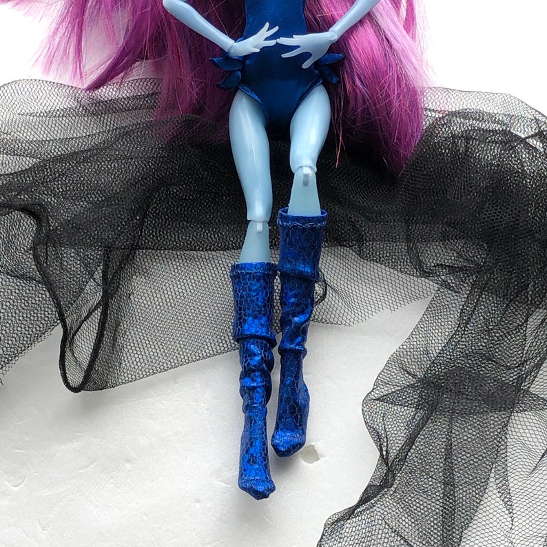 May include: A blue and black doll with long purple hair wearing a blue one-piece swimsuit and blue boots. The doll is posed with one leg bent and the other leg straight. The doll is sitting on a black tulle fabric.