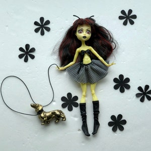 May include: A yellow doll with black and white striped skirt and black boots, holding a leash attached to a gold dachshund figurine. The doll has black hair and red eyes. The background is white with black flower shapes.