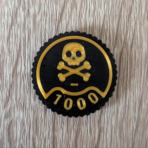 May include: A black and gold circular emblem featuring a skull and crossbones design. The emblem has a scalloped edge and the number "7000" in gold lettering. The background is a light wood grain.