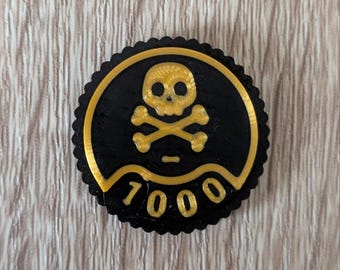 Peut inclure: Un emblème circulaire noir et or avec un motif de tête de mort et d'os croisés. L'emblème a un bord festonné et le chiffre "7000" en lettres dorées. Le fond est un grain de bois clair.