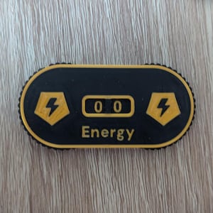 Könnte beinhalten: Schwarzer und gelber Energietracker mit einem digitalen Display, das 00 anzeigt. Der Tracker hat auf beiden Seiten des Displays ein Blitzsymbol und das Wort "Energie" ist unter dem Display aufgedruckt.
