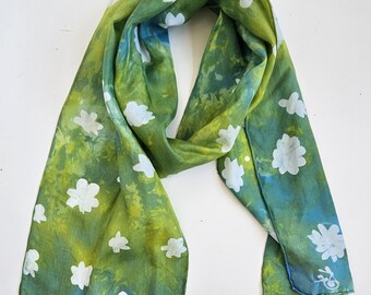 May include: A green and blue silk scarf with a white floral pattern. The scarf is hand-dyed and features a gradient of colors.