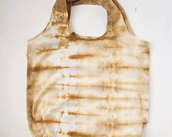 May include: A beige and white tie-dye tote bag with a large handle opening. The bag has a horizontal striped pattern in shades of brown and white. The bag is made of a soft, lightweight fabric.