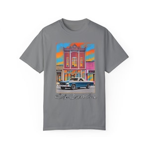 May include: A grey t-shirt featuring a vintage illustration of a blue car driving past a pink building with the text "B.B.M.F.S.A." on the front. The text "E.J. Canabra" is printed below the illustration.