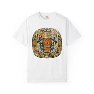 May include: A white short-sleeve t-shirt featuring a large graphic of a gold championship ring. The ring design includes the text NEW YORK PHISH 2026, a basketball icon, the number 5, and the words SHOWS AT THE GARDEN CHAMPIONS in gold and blue.