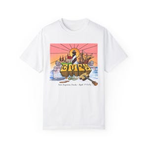 May include: White t-shirt with a colourful graphic design featuring a lighthouse, a ship, and the text "GMES" with the words "Saint Augustine, Florida - April 3'3 2023" below.
