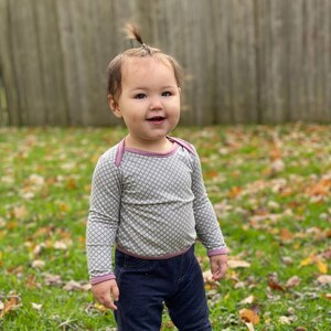 May include: A young child wearing a grey and white patterned long-sleeved shirt with a pink trim and dark blue jeans stands in a grassy area with fallen leaves.