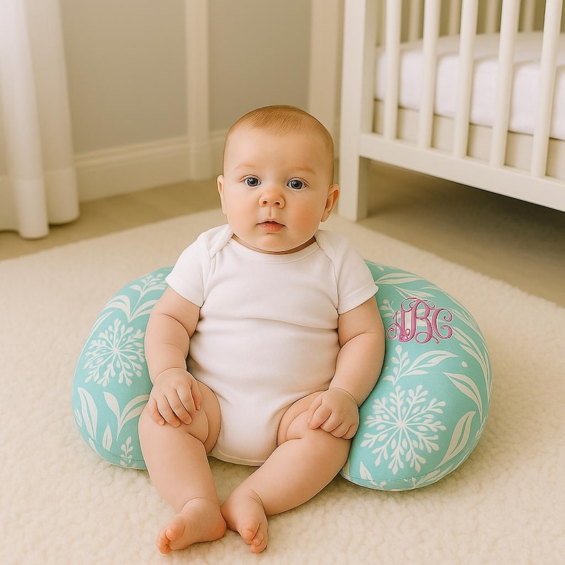 May include: A baby sits on a turquoise nursing pillow with a white floral design and a pink monogram. The pillow is crescent-shaped and rests on a white rug. The baby wears a white onesie.