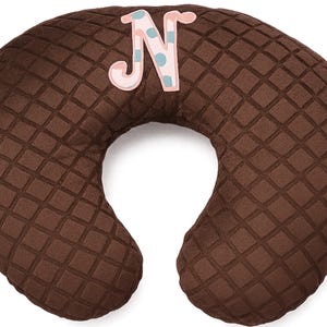 May include: A brown, U-shaped nursing pillow with a diamond pattern. A decorative letter "N" in pink and blue polka dots is attached to the pillow. The pillow is designed for comfort and support.