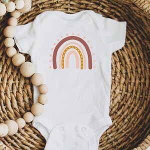 May include: A white baby onesie with a pink, brown, and orange rainbow graphic. The rainbow has a pattern of small hearts and dots.