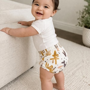 May include: A smiling baby wearing a white onesie and patterned diaper cover stands on a white rug. The diaper cover features a cream background with brown and gold cactus and abstract shapes.