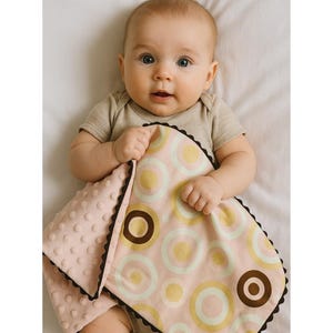 May include: A baby blanket featuring a pink, brown, and yellow circular design. The blanket has a soft, textured pink reverse and a dark brown scalloped edge. The baby is wearing a light brown onesie.
