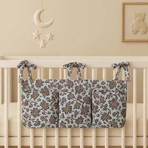 Peut inclure: Un lit de bébé avec un organiseur à motifs floraux attaché sur le côté. L'organisateur a trois poches et est décoré de fleurs marron sur fond bleu clair. Un mobile avec une lune et des étoiles est suspendu au-dessus du lit.